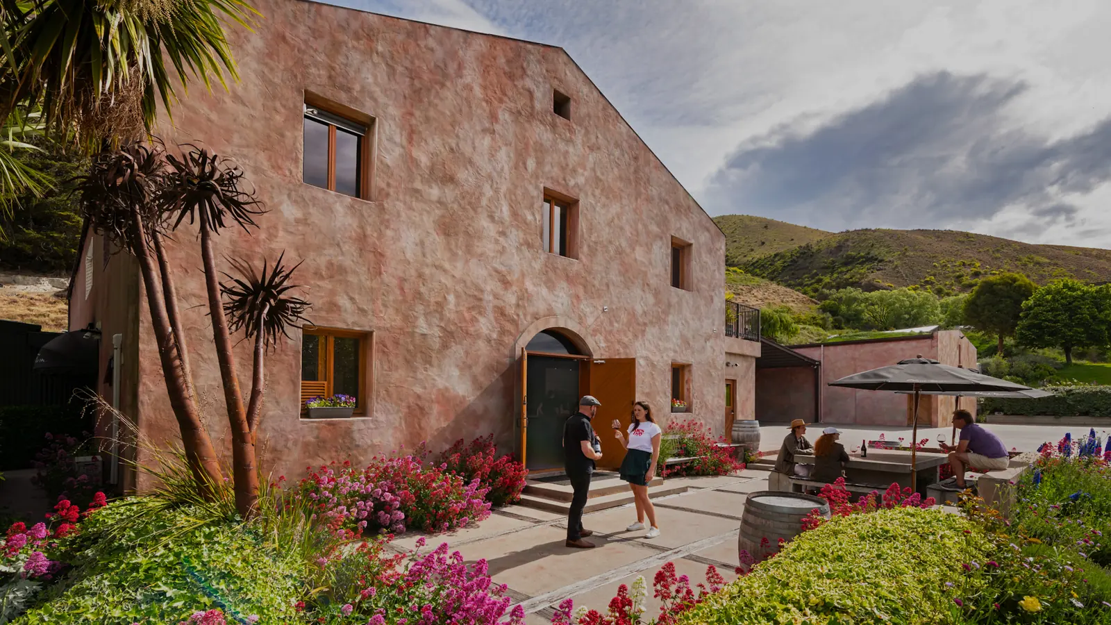 Wine glasses in hand outside the Chard Farm cellar door, guests at tables under an umbrella and dry hills rising behind