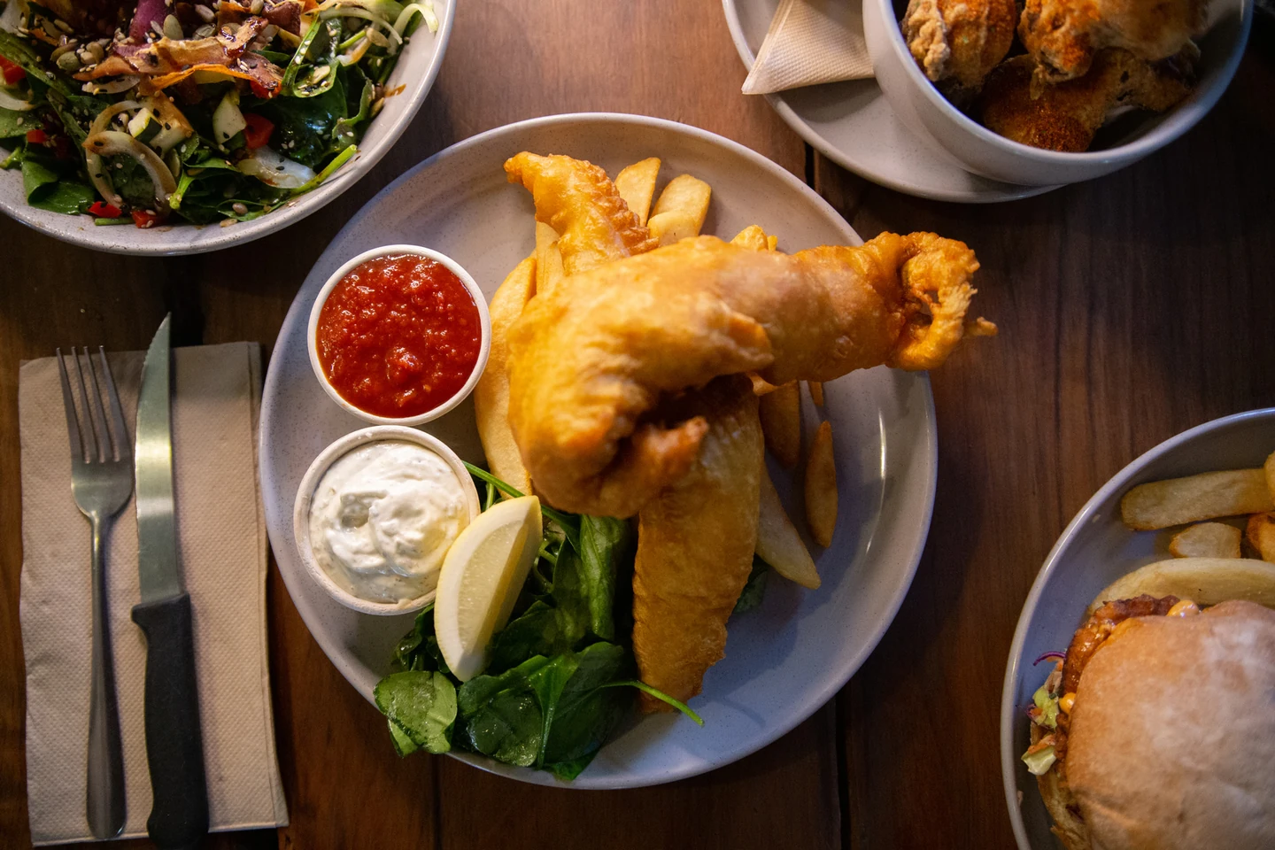 Beer-battered fish and chips with tartare and tomato sauce, a burger and a green salad spread across a dark timber table