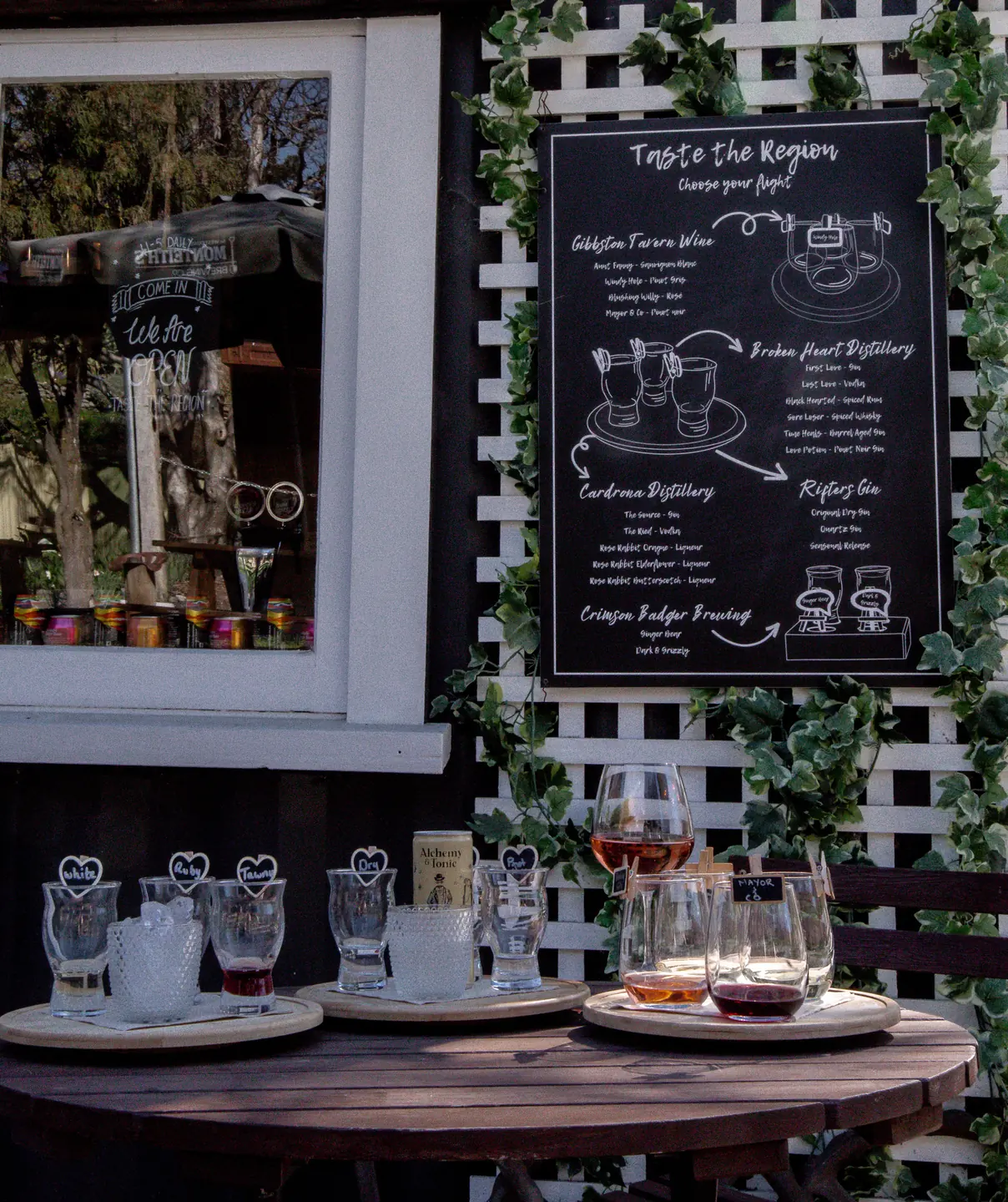 Tasting flights and wine glasses on a round table in the garden, beside a chalkboard listing the Central Otago distilleries poured here