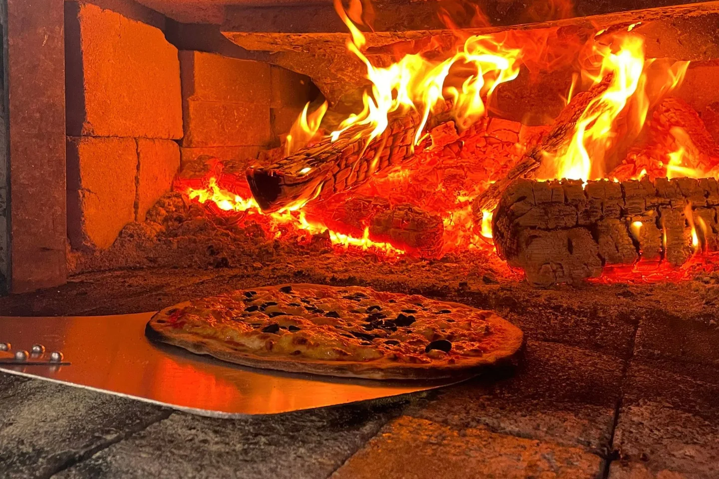 A pizza on a steel peel at the mouth of a brick wood-fired oven, flames burning along the back wall