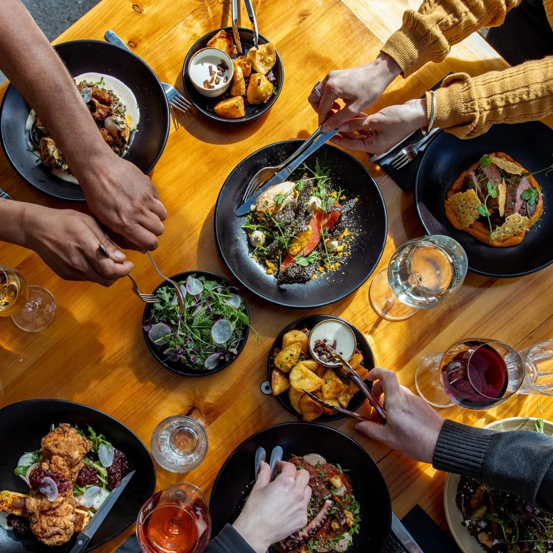Looking down on a bistro table where several people reach across black bowls of shared plates, glasses of rose and red between them