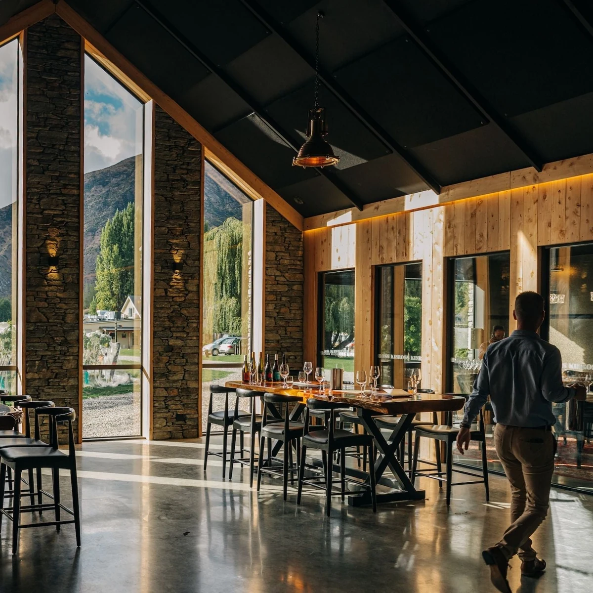 The tasting room's long communal tables and bar stools under a raked black ceiling, floor-to-ceiling windows opening onto the vines