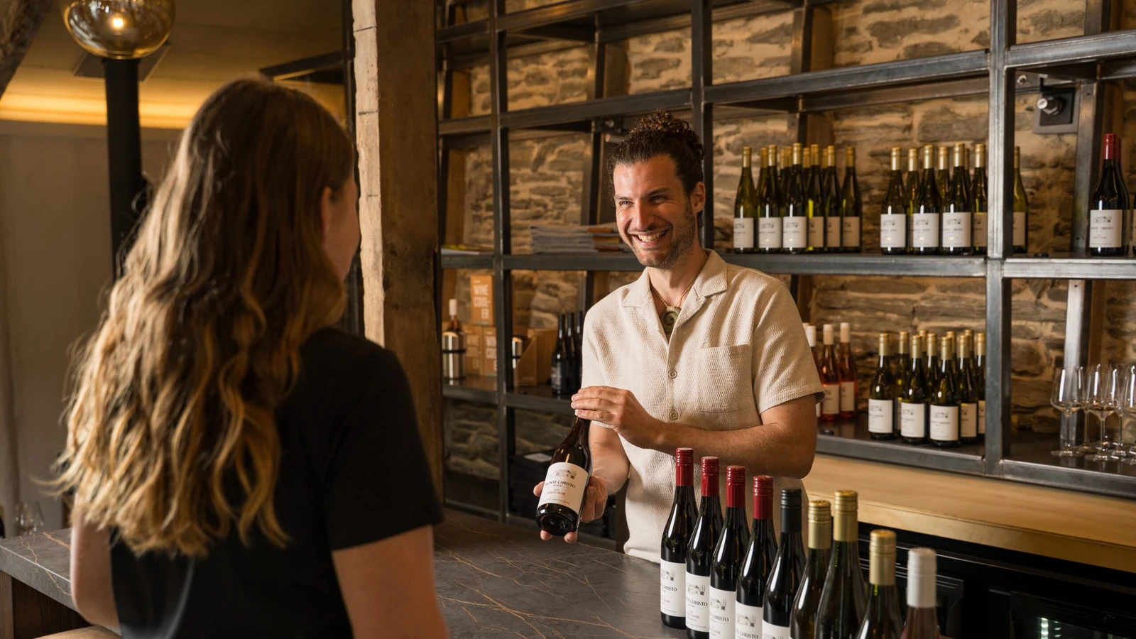 Someone behind the stone bar holding up a bottle to a visitor, a row of reds waiting on the counter and shelves of wine against the stonework