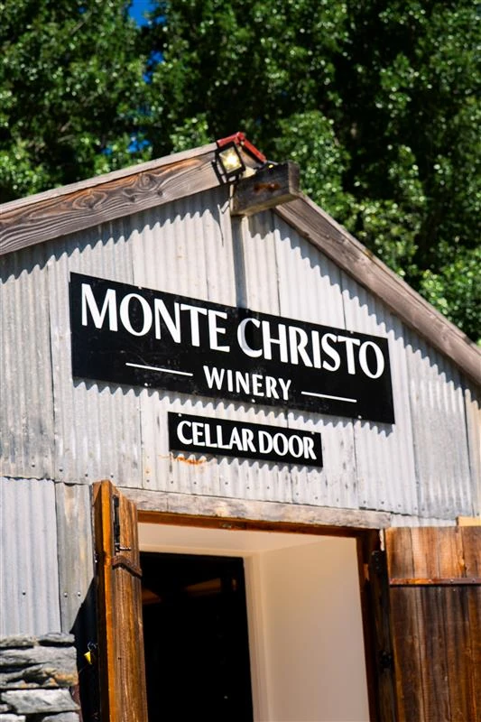 Corrugated iron gable above a stone wall, painted signs reading Monte Christo Winery and Cellar Door, timber doors swung open below