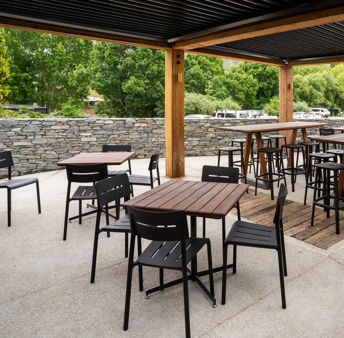 Timber-framed roof over a concrete terrace of dark metal chairs and hardwood tables, a low schist wall along the edge and green willows beyond