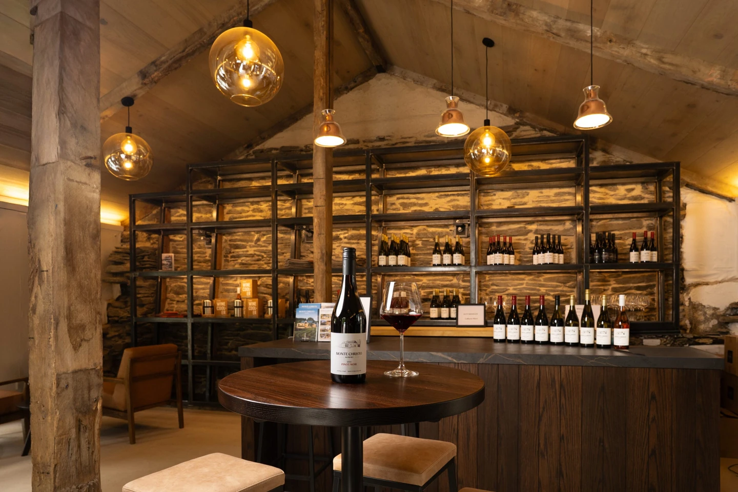 Tasting room under a pitched timber ceiling, dry stone walls behind steel shelving lined with bottles, glass pendant lights over a small round table