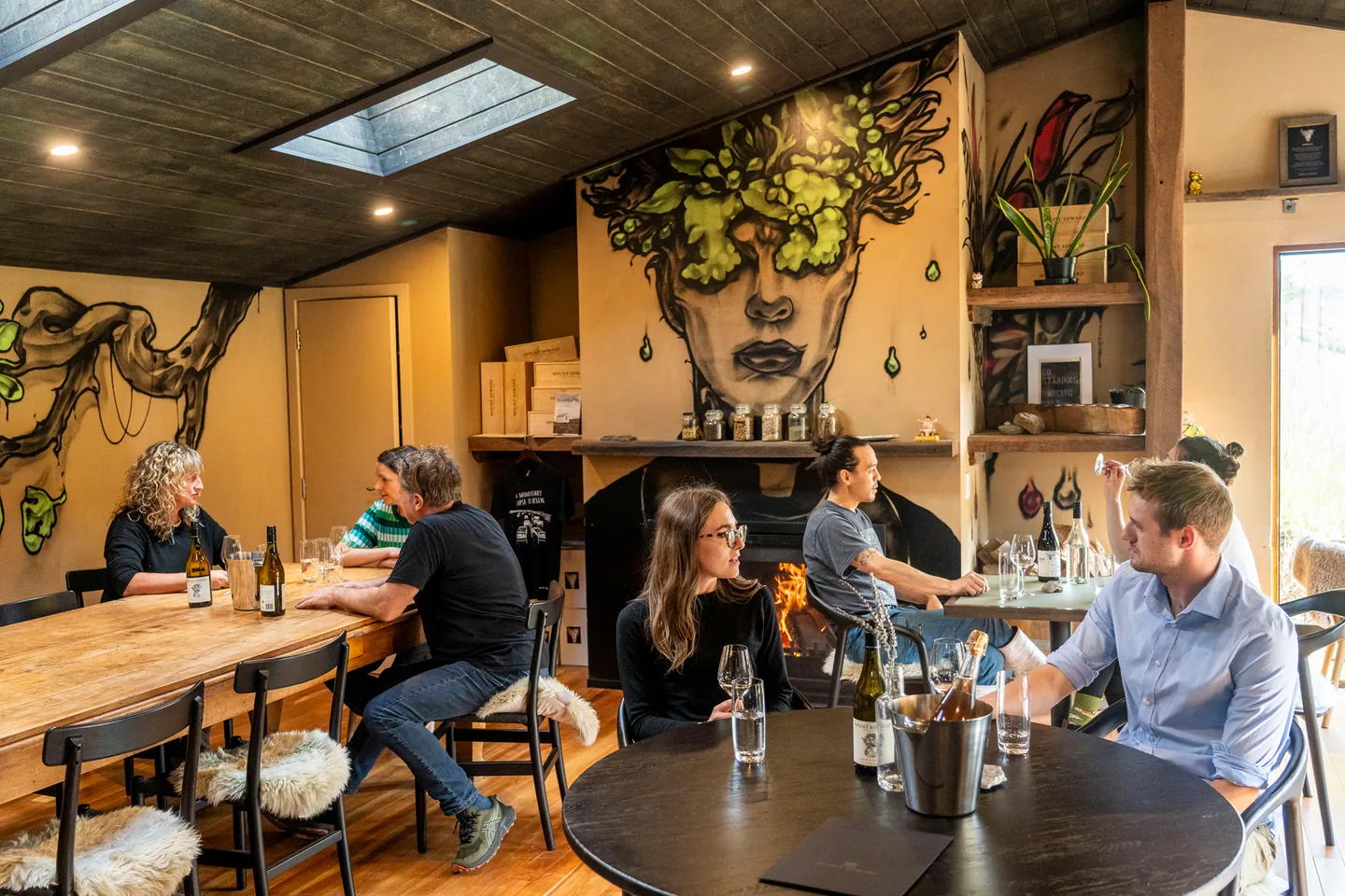 Guests seated at two wooden tables in a small skylit tasting room, bottles and ice buckets between them and a painted mural covering the back wall