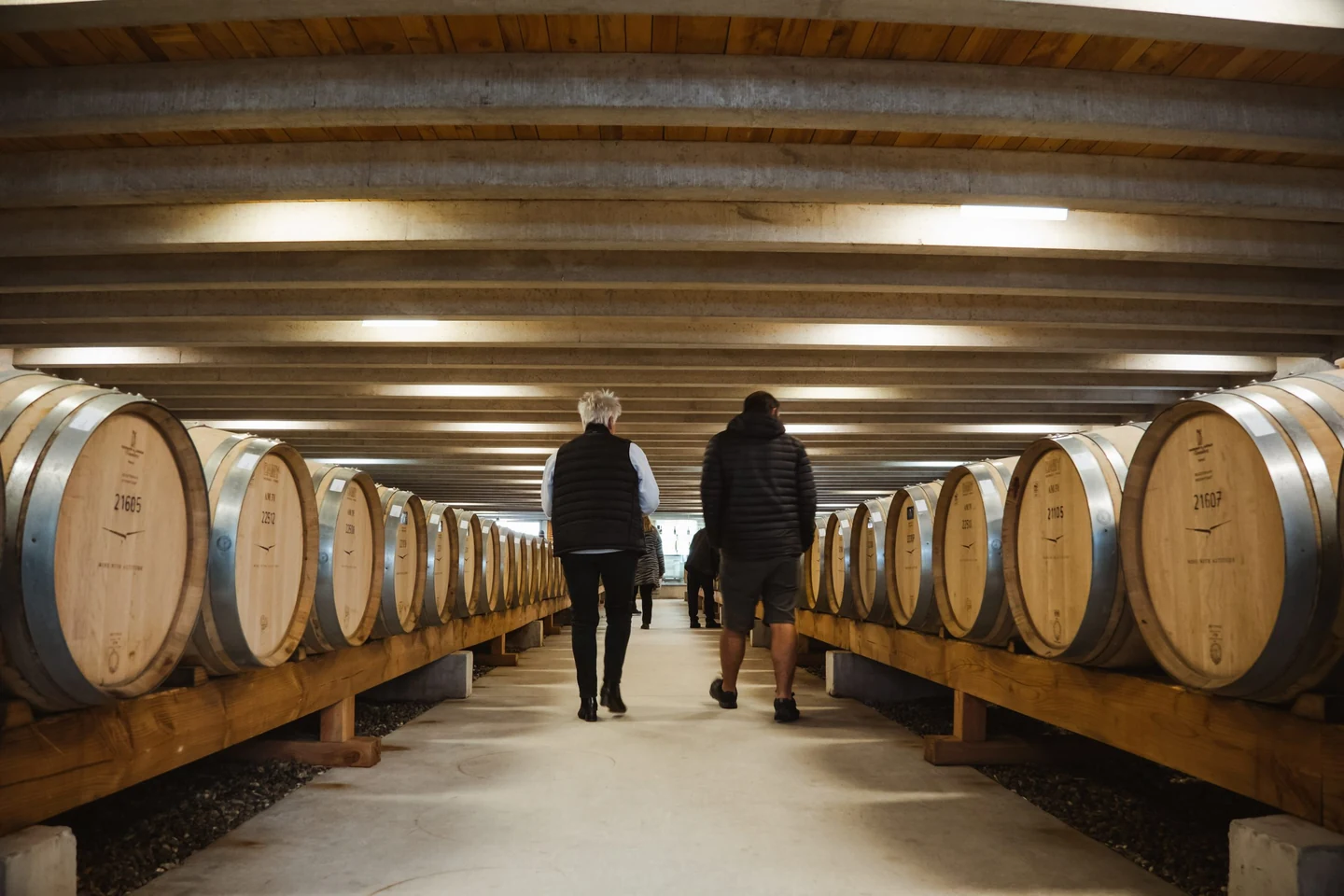 Two visitors walking a long underground hall lined either side with oak barrels beneath a curved timber ceiling