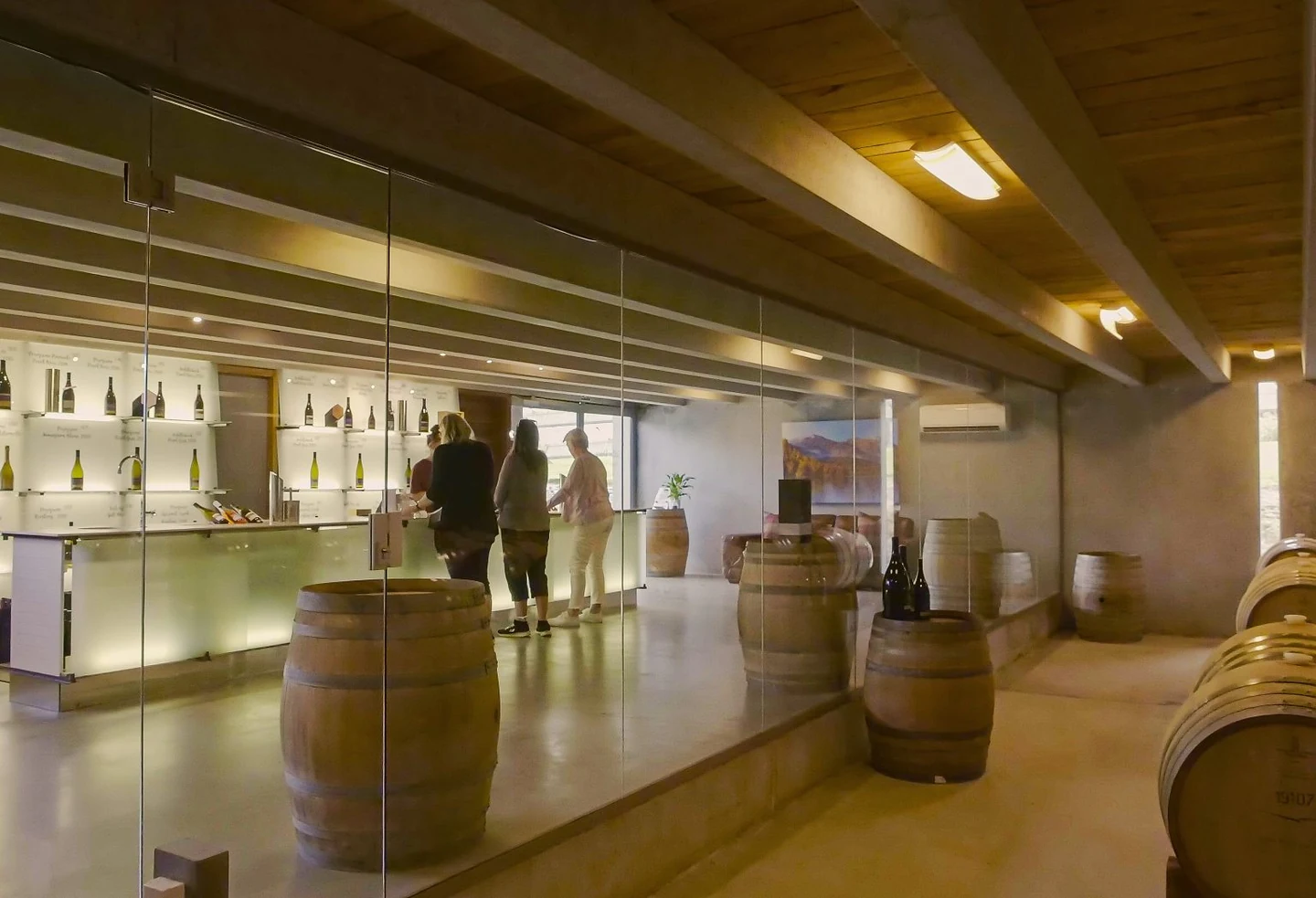Guests at the lit tasting counter behind a glass wall, bottles displayed in backlit alcoves and oak barrels standing on the polished concrete floor