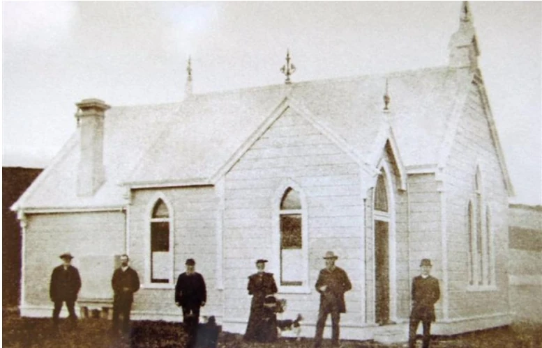 Sepia photograph of six people in Victorian dress standing outside the newly built weatherboard church, with finials and a brick chimney it no longer has