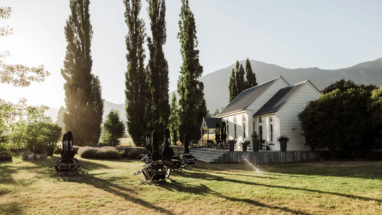 The church at the far end of a mown lawn scattered with folded umbrellas, poplars throwing long shadows across the grass